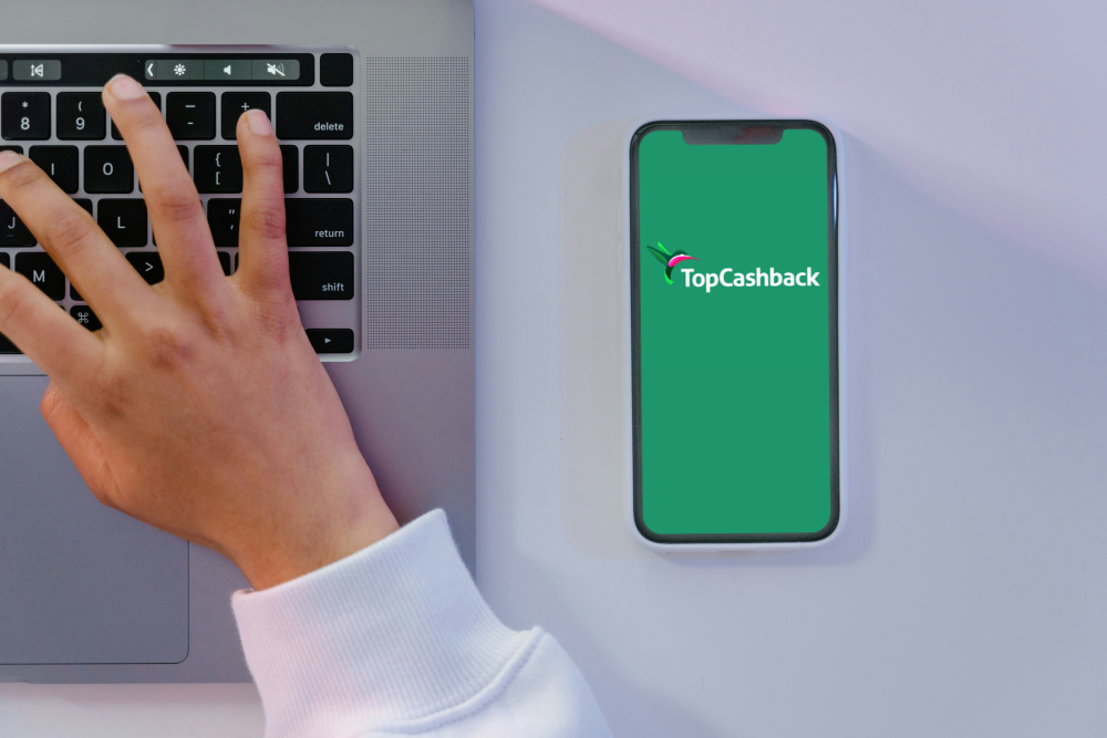 how to use Topcashback 10 ways to maximise your earnings A person’s hand types on a laptop next to a smartphone displaying the TopCashback app logo, showing how to use TopCashback for earning rewards while shopping online.