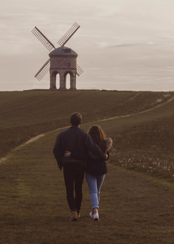 A couple walking through a field with a windmill in the background.
