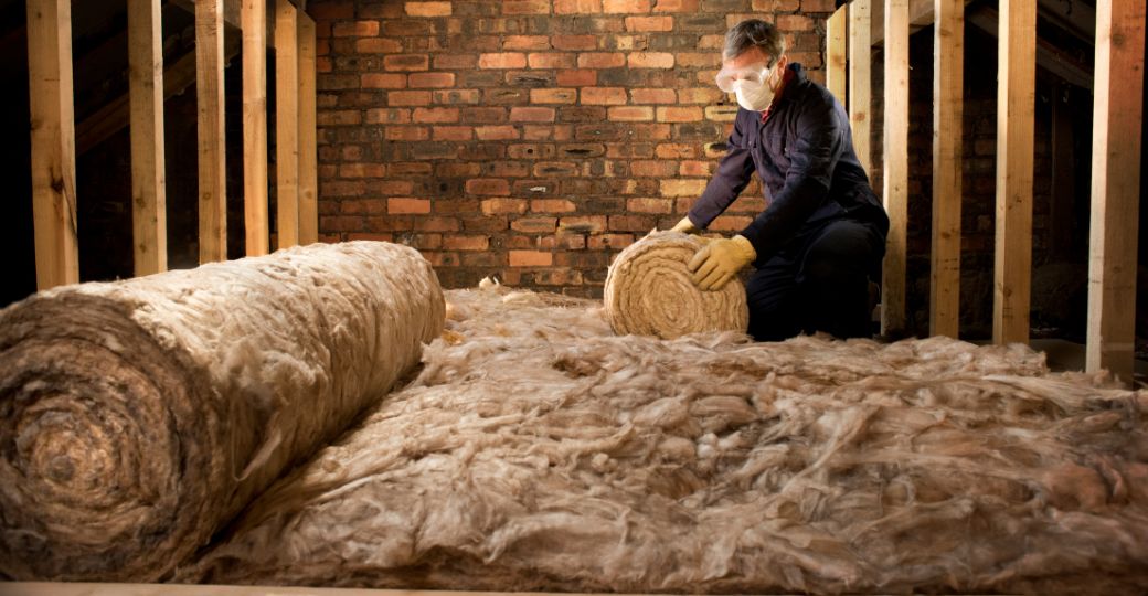 A man is working on a roll of insulation in an attic.