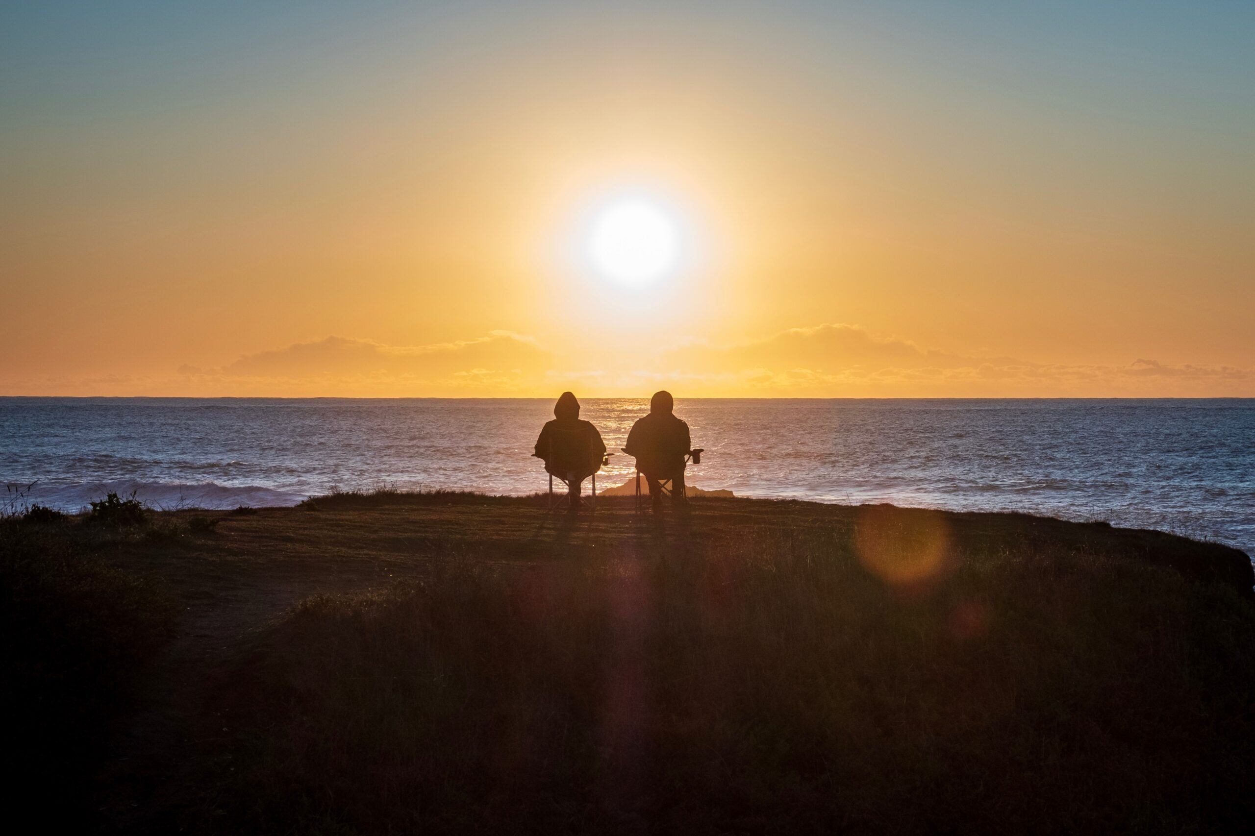 Two people standing on a hill overlooking the ocean at sunset.