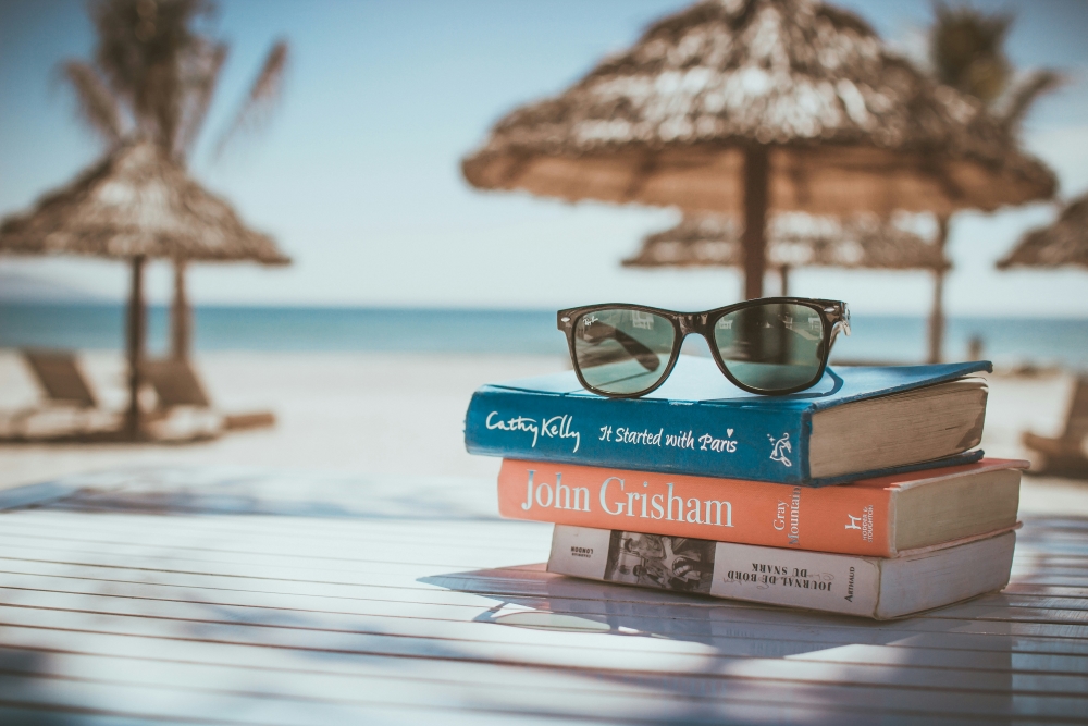A stack of books with sunglasses on top, set on a wooden table at a beach, with straw umbrellas and a clear sky in the background.