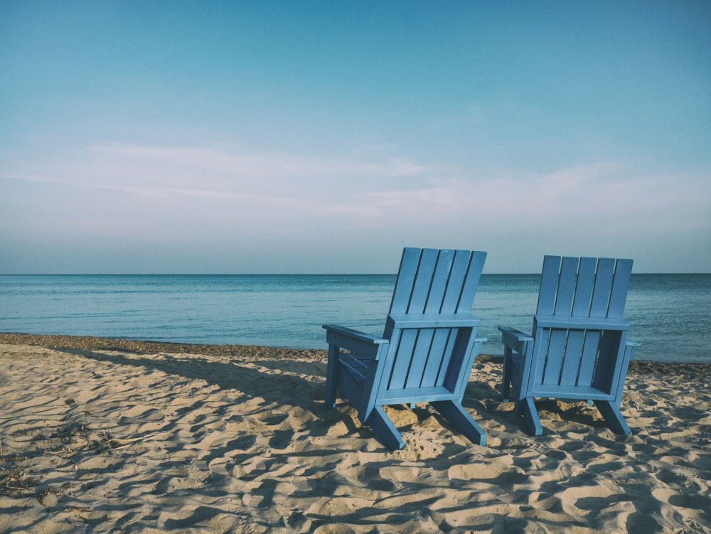 Two blue adirondack chairs facing the ocean on a sandy beach under a clear sky.