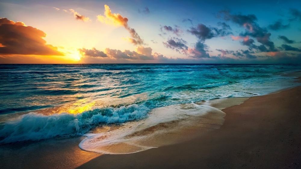 Sunset over a beach with waves gently crashing onto the shore and vibrant clouds in the sky.