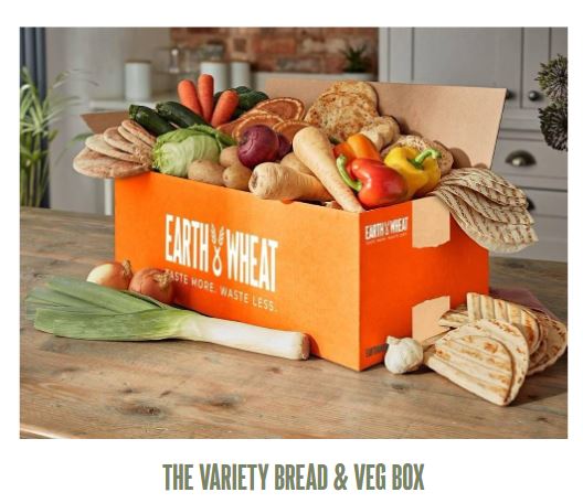 earth and wheat bread box 1 An orange box labeled "Earth & Wheat" filled with assorted breads and vegetables, including peppers, leeks, and various loaves, sits on a wooden table.