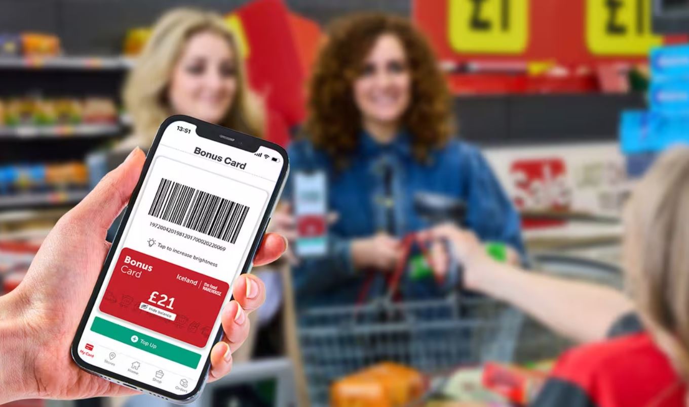 iceland-bonus-card A person holds a smartphone displaying a digital bonus card with a £21 balance in a supermarket while two women check out groceries in the background.