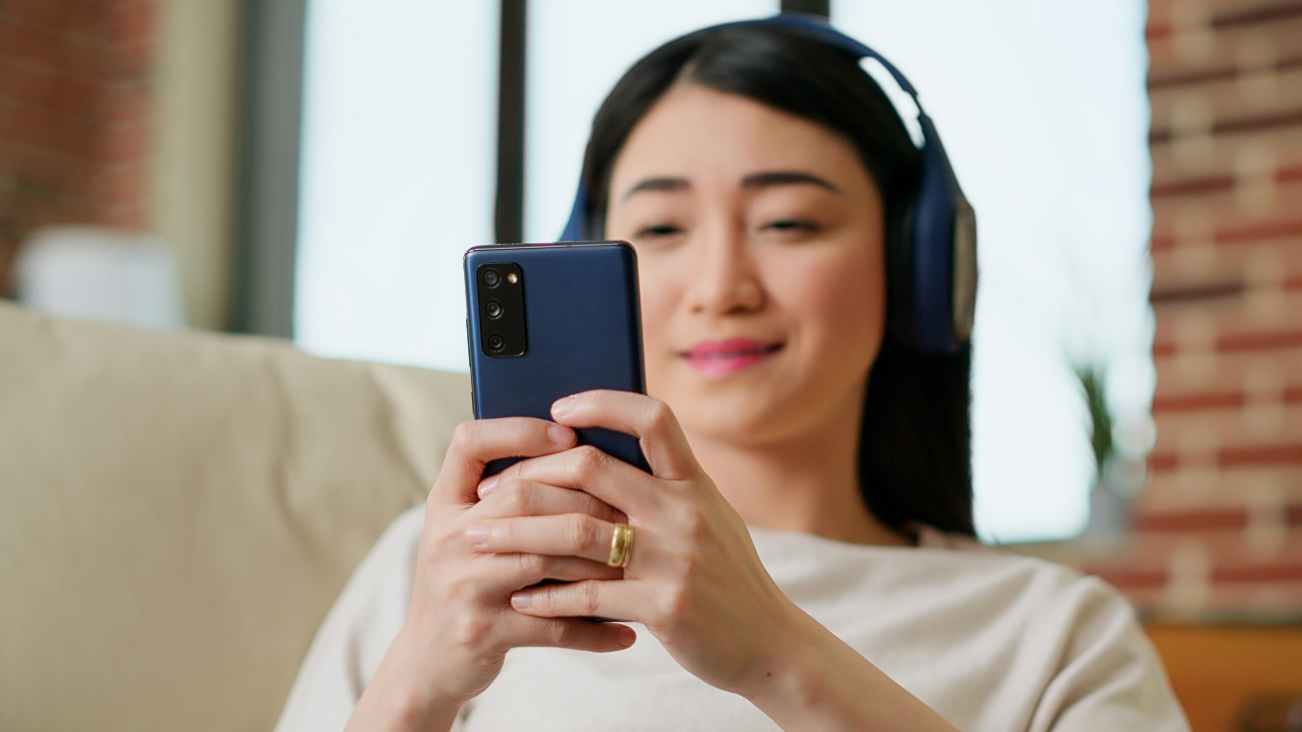 mystery shopper unsplash 2 A woman wearing headphones smiles while using a smartphone, sitting indoors on a light-colored couch.