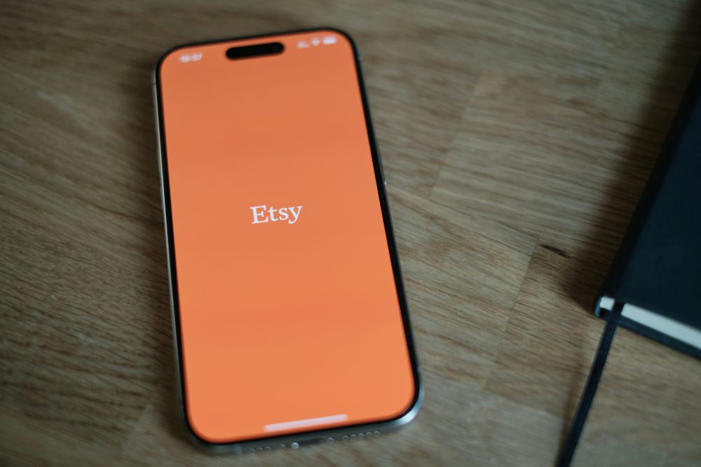 A smartphone displaying the orange Etsy app welcome screen rests on a light wooden surface next to a closed black notebook.