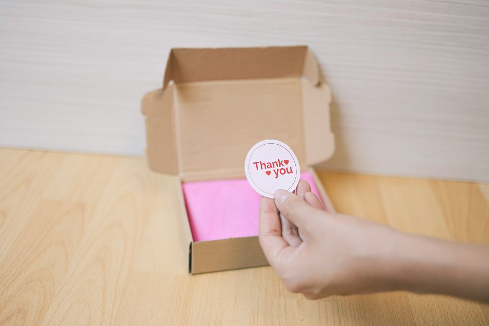A hand holds a round "Thank you" sticker above an open cardboard box with pink tissue paper inside, on a wooden surface.