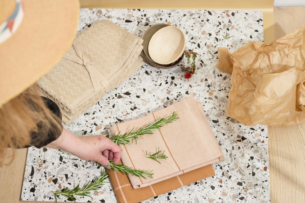 Person arranging folded towels and sprigs of rosemary on a speckled surface, with a small bowl, flower, and crumpled brown paper nearby.