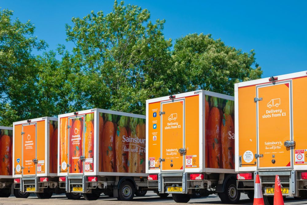 Sainsburys vans Four Sainsbury’s delivery trucks with orange branding are parked in a row outdoors, with trees and a clear blue sky in the background.