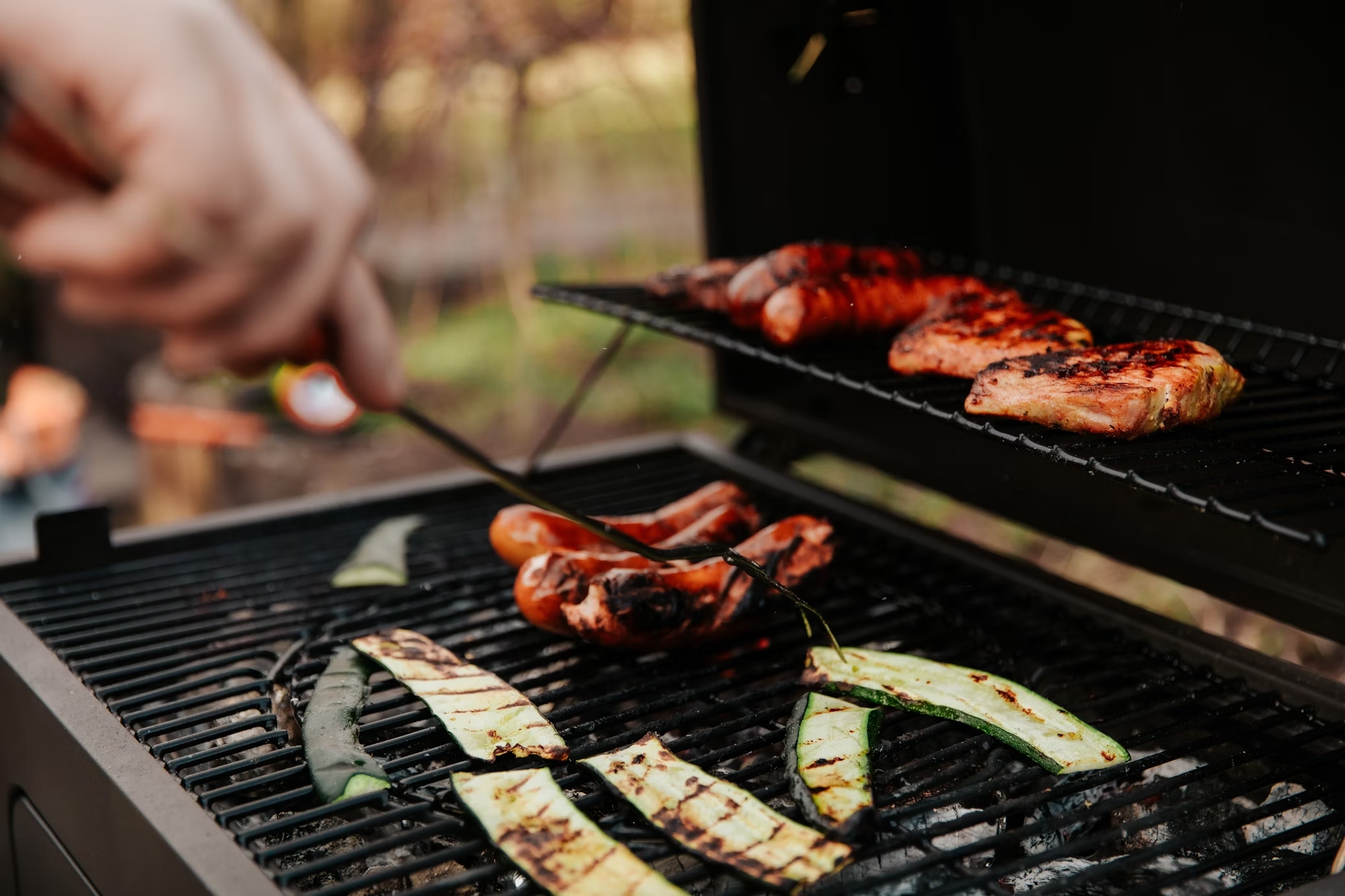 Sausages on barbecue A person grills sausages, salmon, and sliced zucchini on a two-level barbecue grill outdoors.