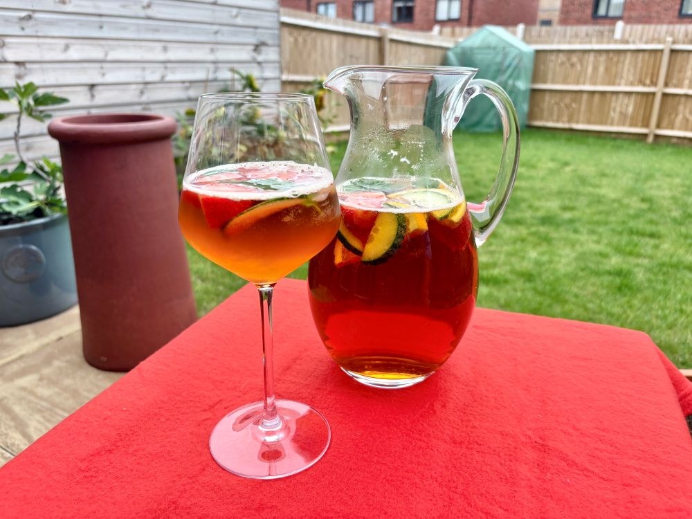A glass and a pitcher of iced tea with lemon and fruit slices sit on a red tablecloth in a backyard setting.