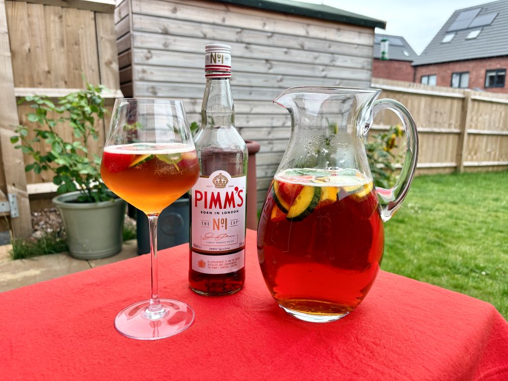 A glass, a bottle of Pimm's No.1, and a pitcher of Pimm's with fruit slices are placed on a red tablecloth in a garden setting.