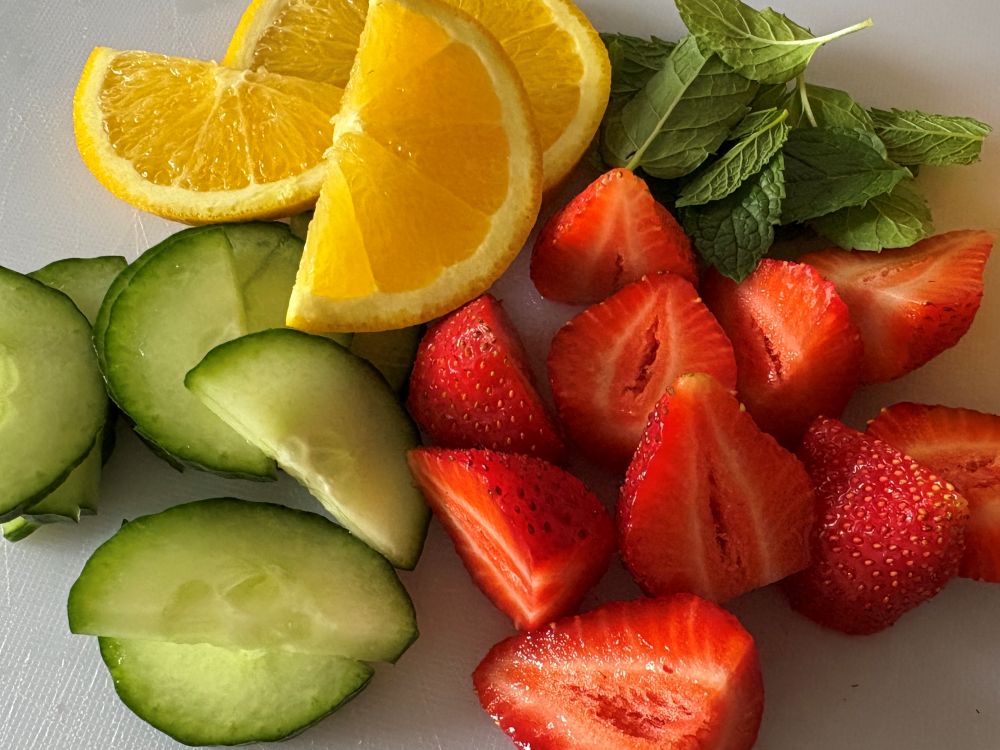 Sliced orange, cucumber, and strawberries arranged next to fresh mint leaves on a white surface.