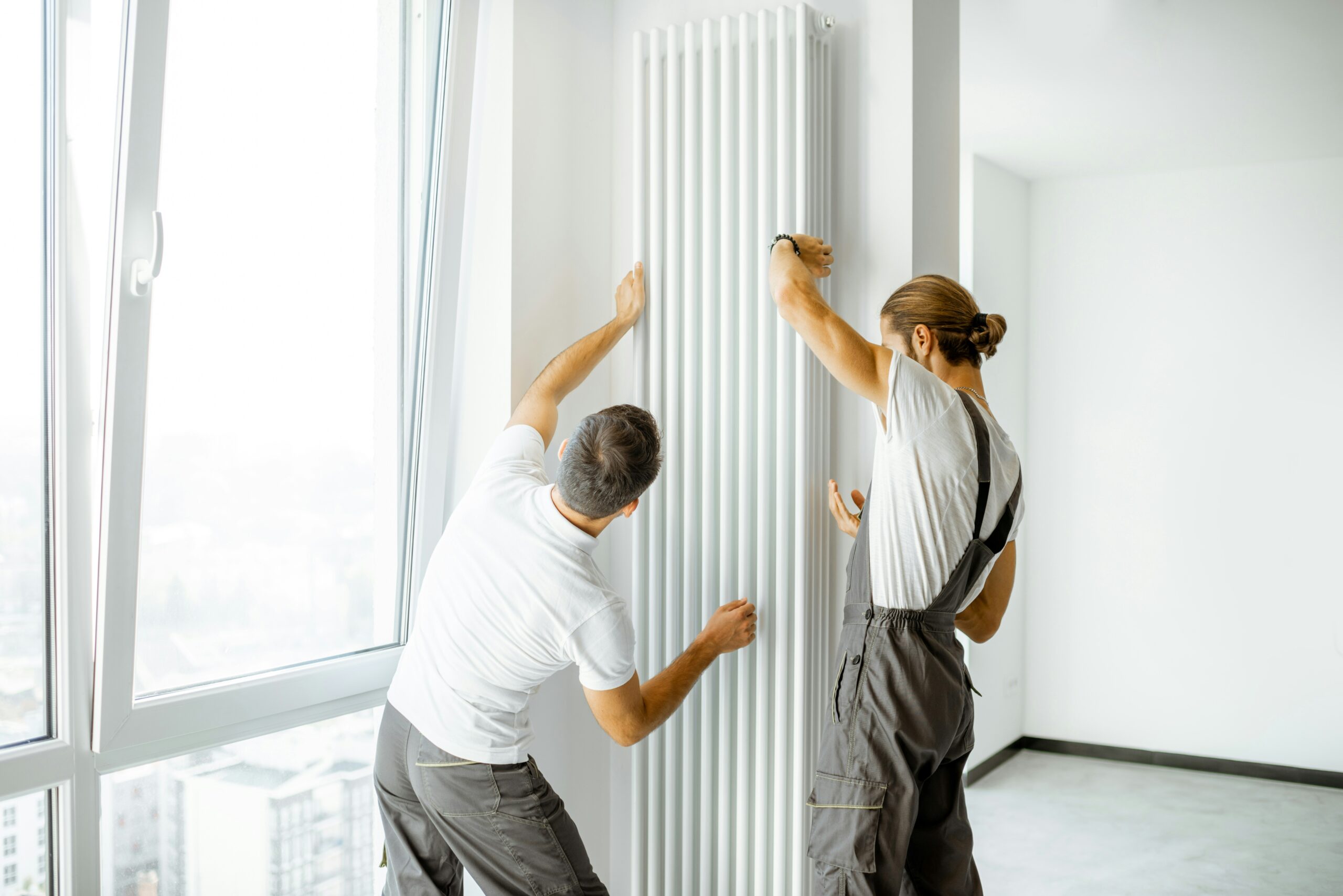 Two workers in overalls install a tall, white vertical radiator on the wall of a bright, modern room with large windows.