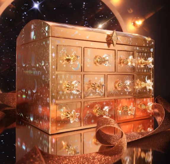 A gold and decorated advent calendar box with twelve small drawers, jeweled knobs, and star, moon, and celestial designs, placed on a ribbon against a sparkly background.