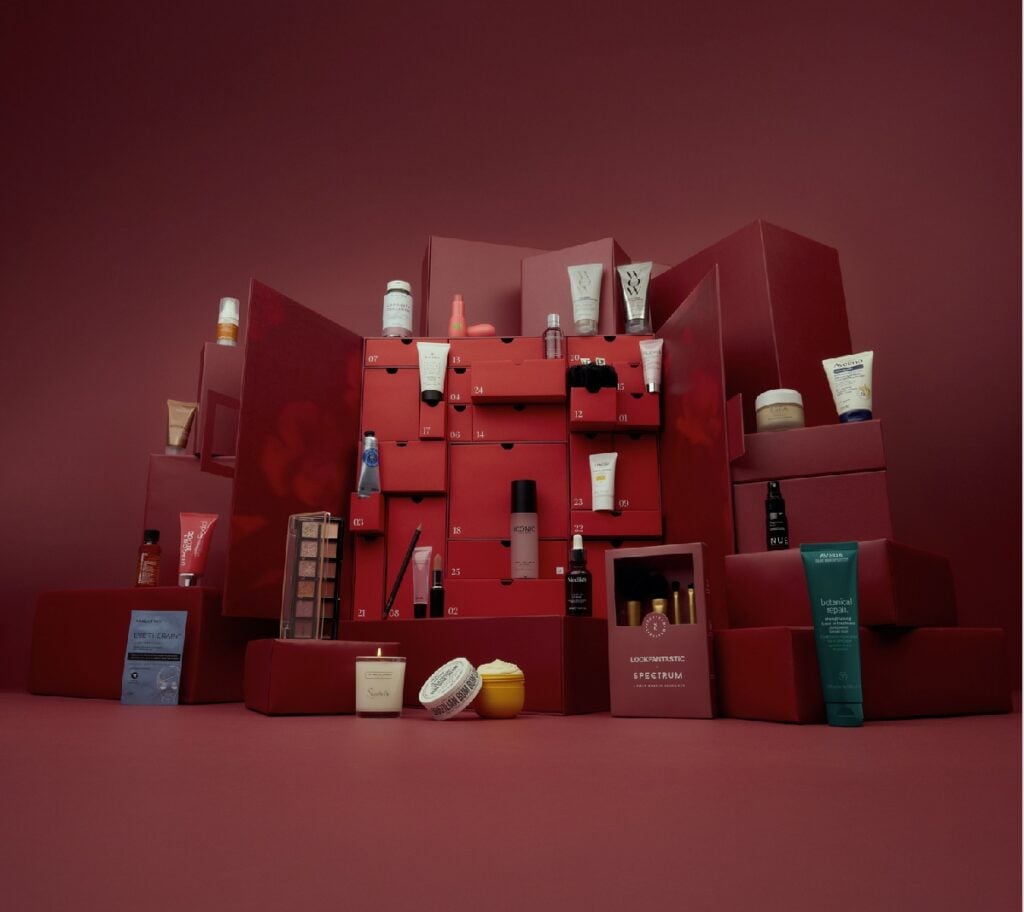 A red advent calendar box with open compartments displays various skincare and beauty products against a matching red background.
