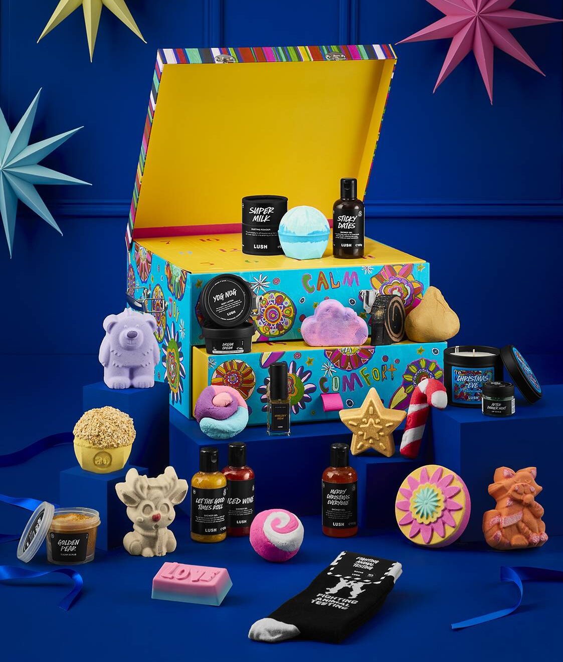 A colorful gift box with “Calm & Comfort” text is open, displaying various bath and body products, soap bars, and festive decorations against a blue background with paper stars and garlands.