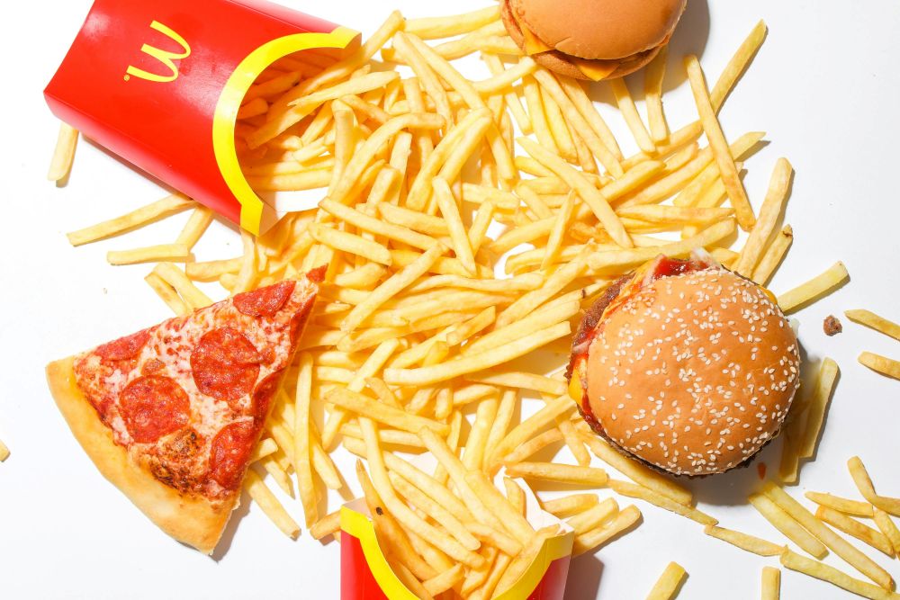 McDonalds food French fries scattered on a white surface with two empty McDonald's fry containers, a pepperoni pizza slice, a cheeseburger, and a hamburger.