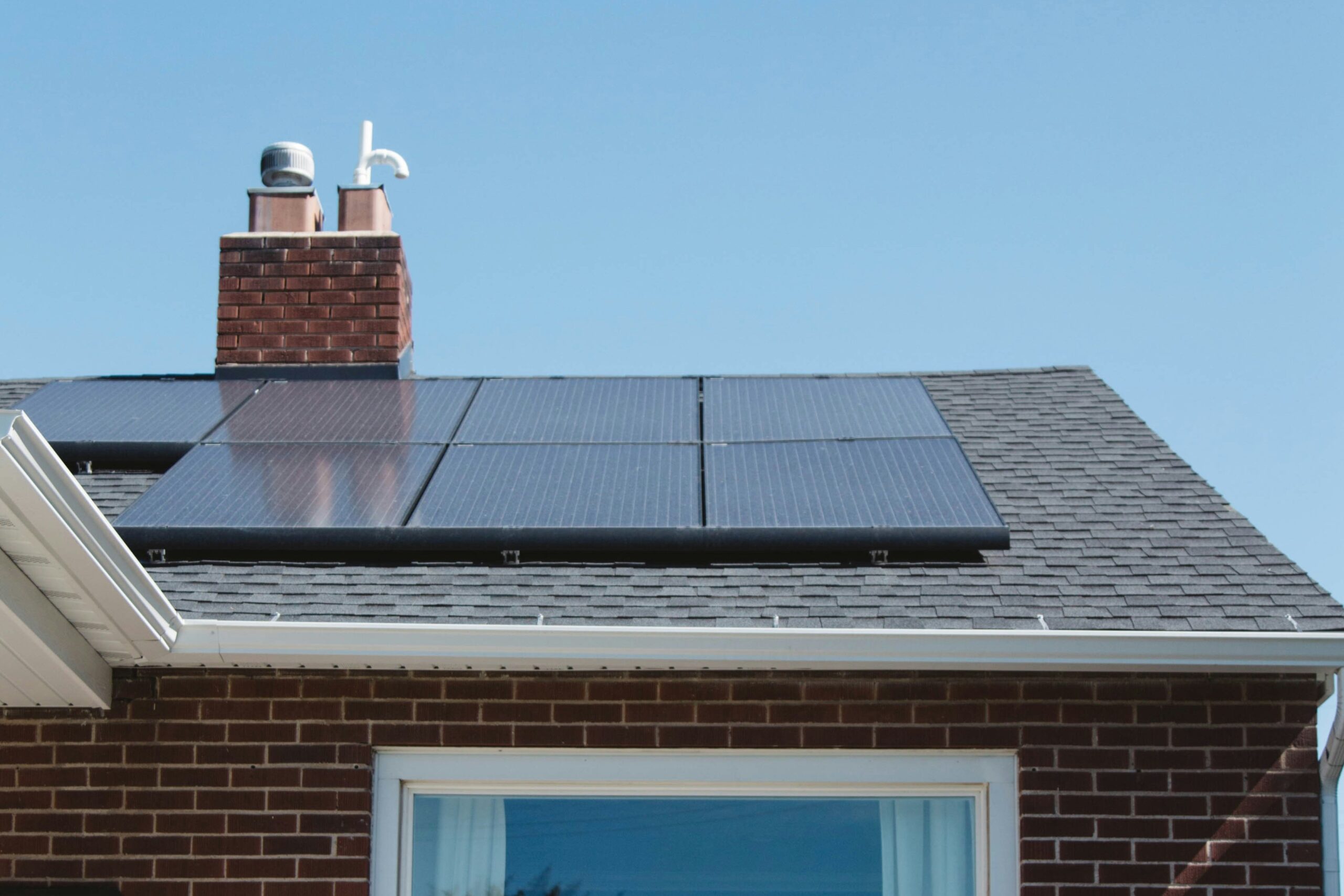 Monocrystalline panels Black solar panels are installed on the roof of a brick house with a chimney, under a clear blue sky.