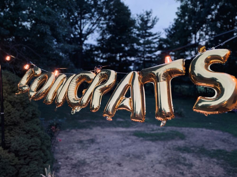 Gold foil balloons spelling "CONGRATS" are strung outdoors with lights, marking a Brits-style promotion against trees and a dirt ground at dusk.