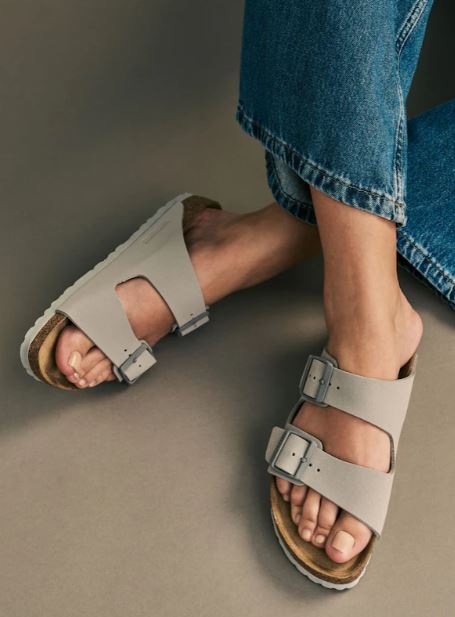 A person wearing light gray double-strap sandals with buckles and blue jeans, standing on a neutral background.