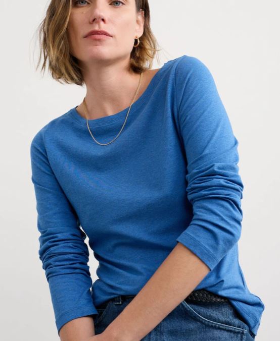 Woman with short brown hair wearing a blue long-sleeve shirt and gold necklace, seated against a plain light background.