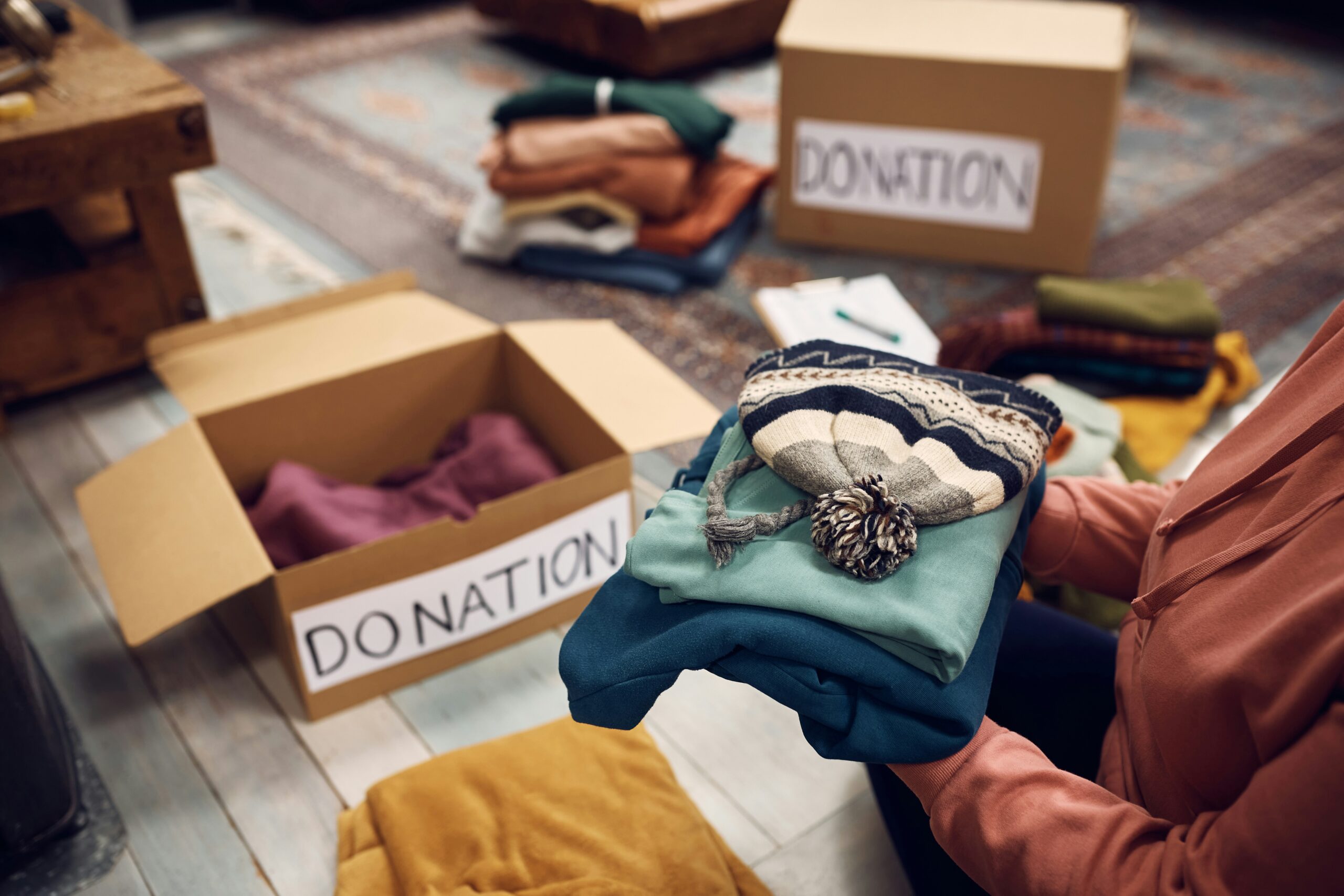 Person holding folded clothes near open donation boxes labeled "donation," with more clothes and boxes in the background, organising a capsule wardrobe
