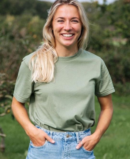 A woman with long blonde hair in a green t-shirt and blue jeans stands outdoors, smiling with her hands in her pockets.