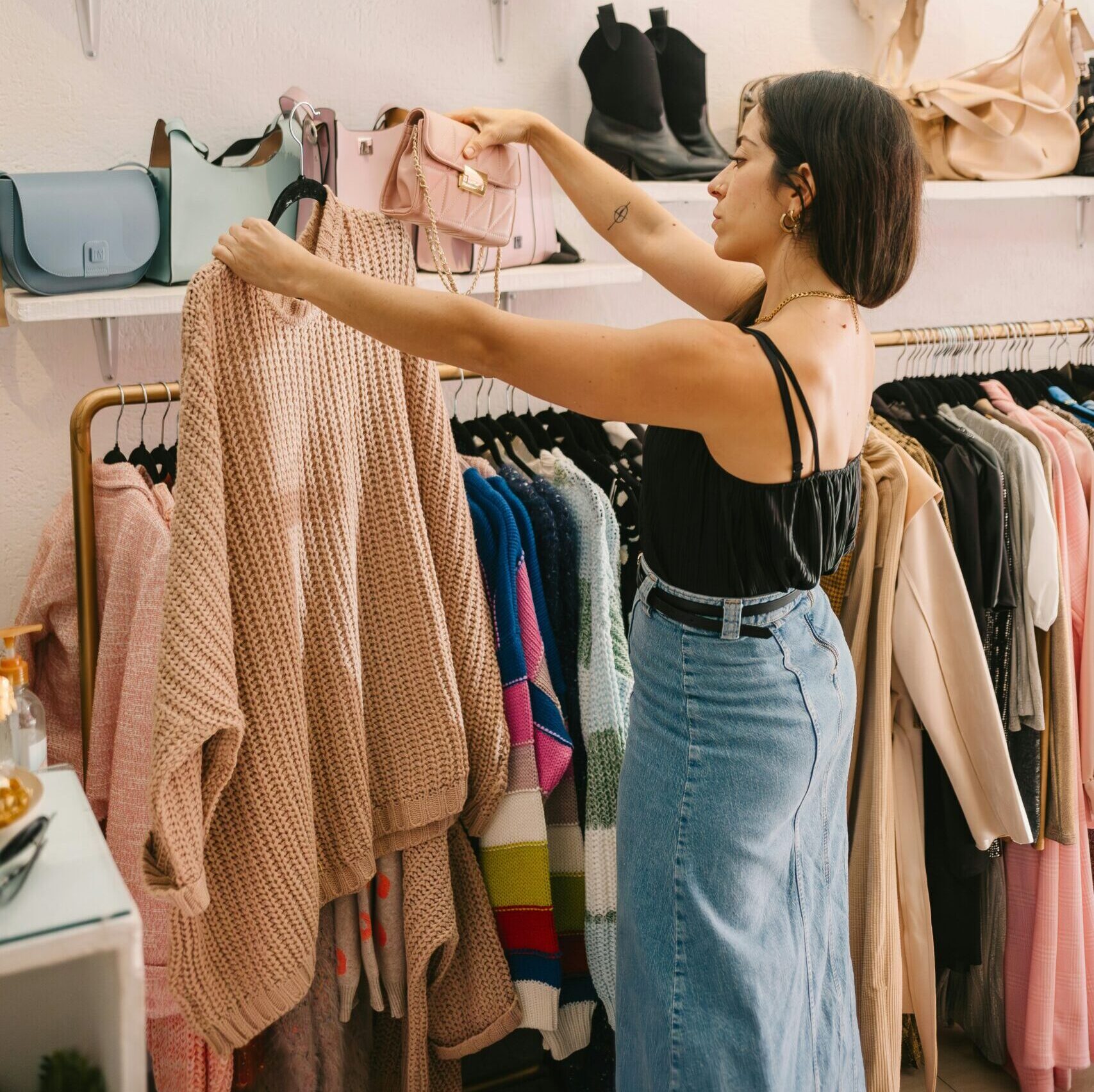 A woman in a black top and denim skirt shops in a boutique, holding a pink handbag and examining a beige sweater among clothing and handbags on display.