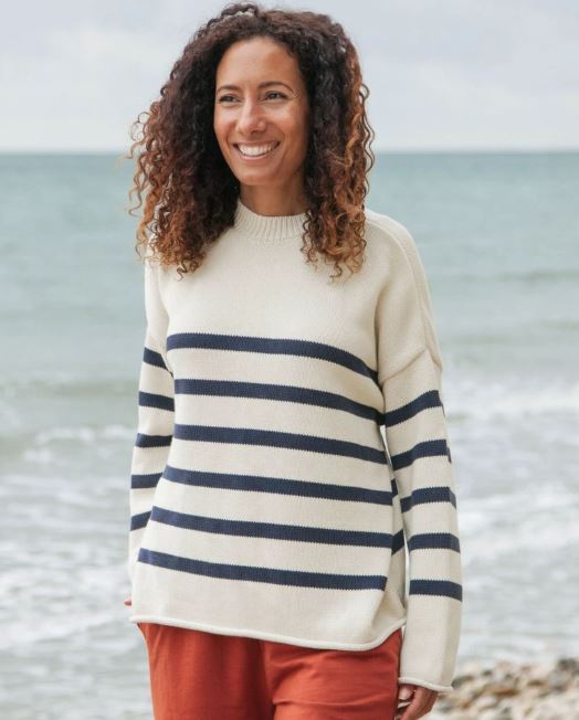 A woman with curly hair stands on a beach, smiling, wearing a cream-colored sweater with navy stripes and rust-colored pants. The ocean and cloudy sky are in the background.