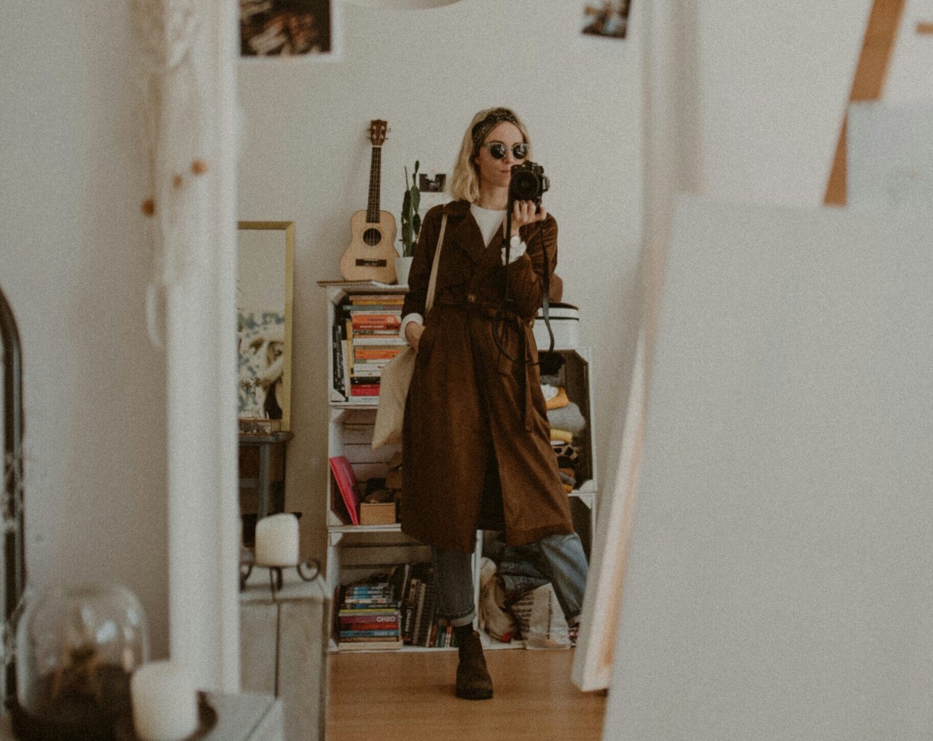 Person taking a mirror selfie of her capsule wardrobe outfit in a cozy, decorated room with art, books, and a guitar visible in the background.