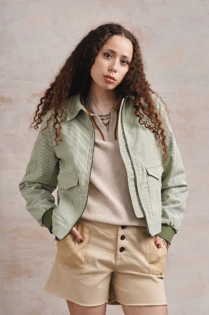 A woman with long curly hair wears a light green jacket, beige top, and tan shorts while standing against a neutral background.