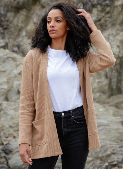 A woman stands outdoors in front of rocks, wearing a beige cardigan over a white t-shirt and black jeans, with one hand touching her hair.