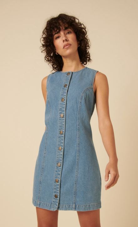 A woman wearing a sleeveless, button-front denim dress stands against a beige background.