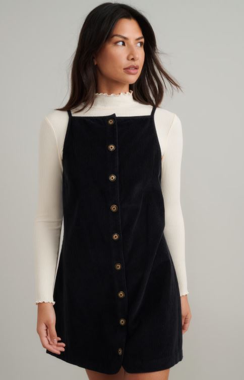 Woman wearing a cream ribbed turtleneck top under a black sleeveless button-down dress, standing against a plain light grey background.