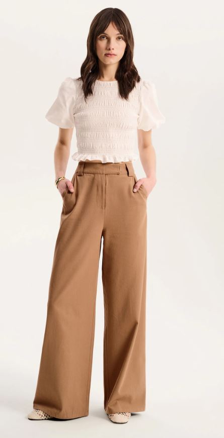 A woman stands with hands in pockets, wearing a short-sleeve white smocked top and high-waisted, wide-leg tan trousers, with beige shoes on a light background.