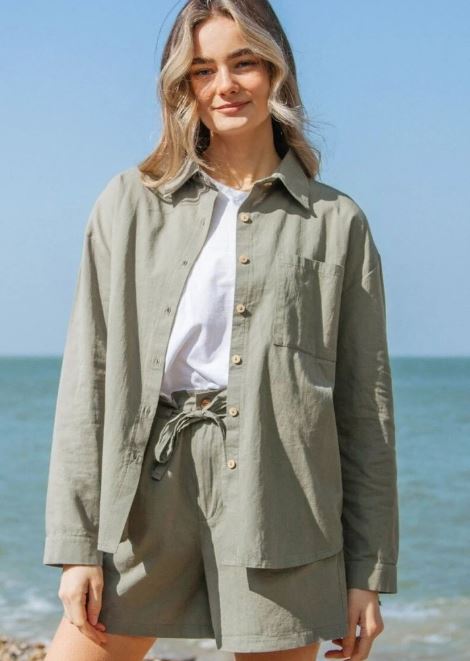 A person stands outdoors by the sea, wearing a light sage green button-up shirt and matching shorts over a white T-shirt.