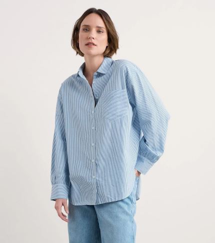 A person wearing a loose-fitting, light blue and white striped button-up shirt with blue jeans stands against a plain light background.