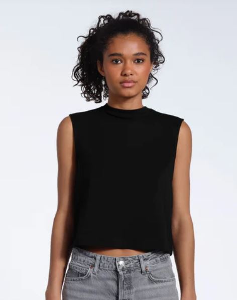 A person with curly hair wearing a sleeveless black top and gray jeans stands against a plain light background.