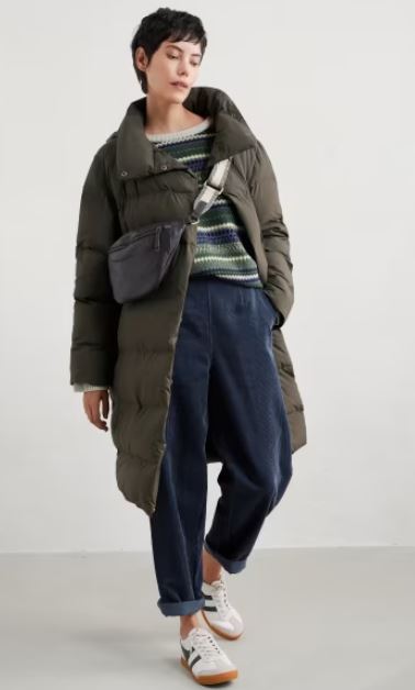 A person wearing a long olive-green puffer coat, striped sweater, navy pants, white sneakers, and a crossbody bag stands against a plain background.