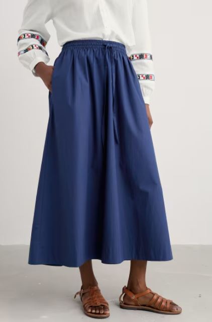 A person wearing a white blouse with patterned sleeve bands, a blue ankle-length skirt with a drawstring waist, and brown sandals, standing against a plain background.