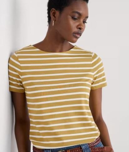 Person wearing a mustard yellow and white striped short-sleeve t-shirt, blue jeans with a brown braided belt, standing against a white wall.