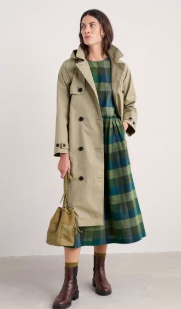 Woman standing in a beige trench coat over a green and blue plaid dress, holding a tan handbag, and wearing brown boots with yellow socks.