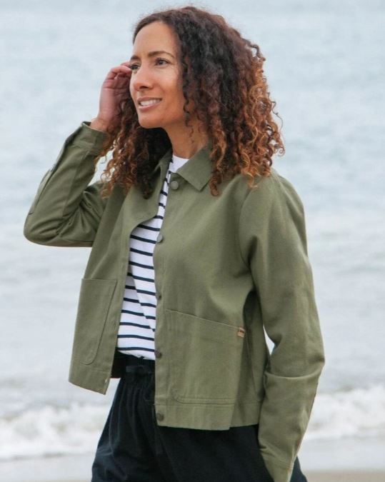 A woman with curly hair stands by the water, wearing a green jacket over a striped shirt and black pants, touching her hair and smiling.