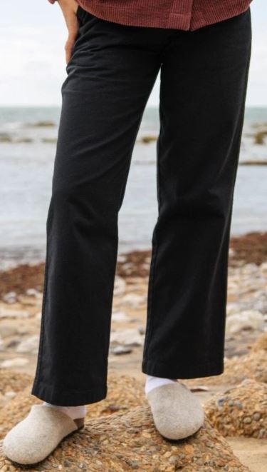 A person stands on a rocky shore wearing black pants, white socks, and beige slippers. The ocean is visible in the background.