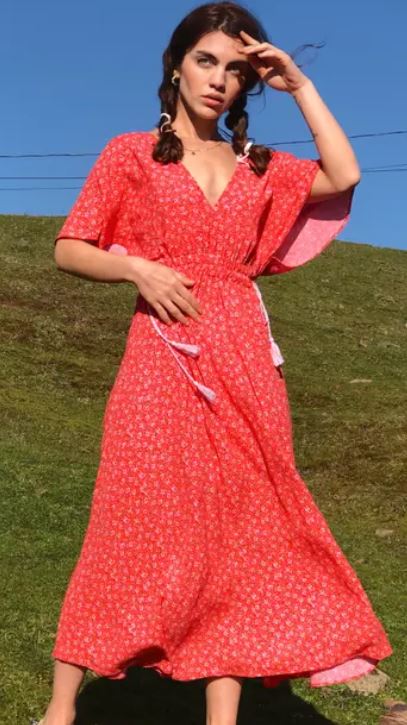 Woman standing outdoors on grass, wearing a long, red floral dress with short sleeves, holding one hand near her head and looking towards the distance.