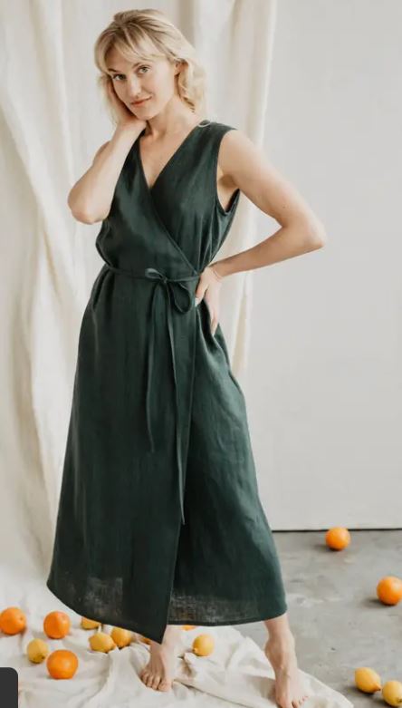 A woman in a sleeveless dark green wrap dress stands barefoot on a light floor with scattered oranges, posing in front of a draped fabric backdrop.