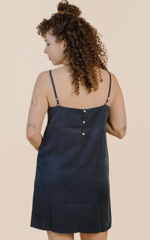 A person with curly hair wears a navy sleeveless dress with thin straps and back button details, standing with their back to the camera.