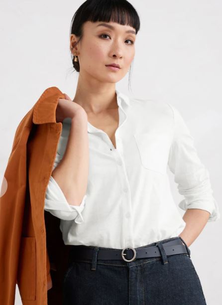 Woman wearing a white button-up shirt and dark jeans, holding an orange jacket over her shoulder, standing against a plain background.
